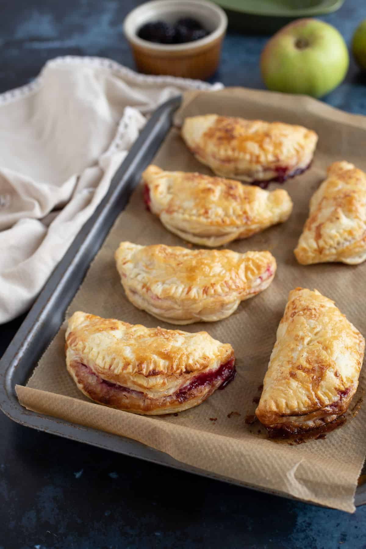 6 apple and blackberry turnovers on a baking tray with cooking apples in the background.