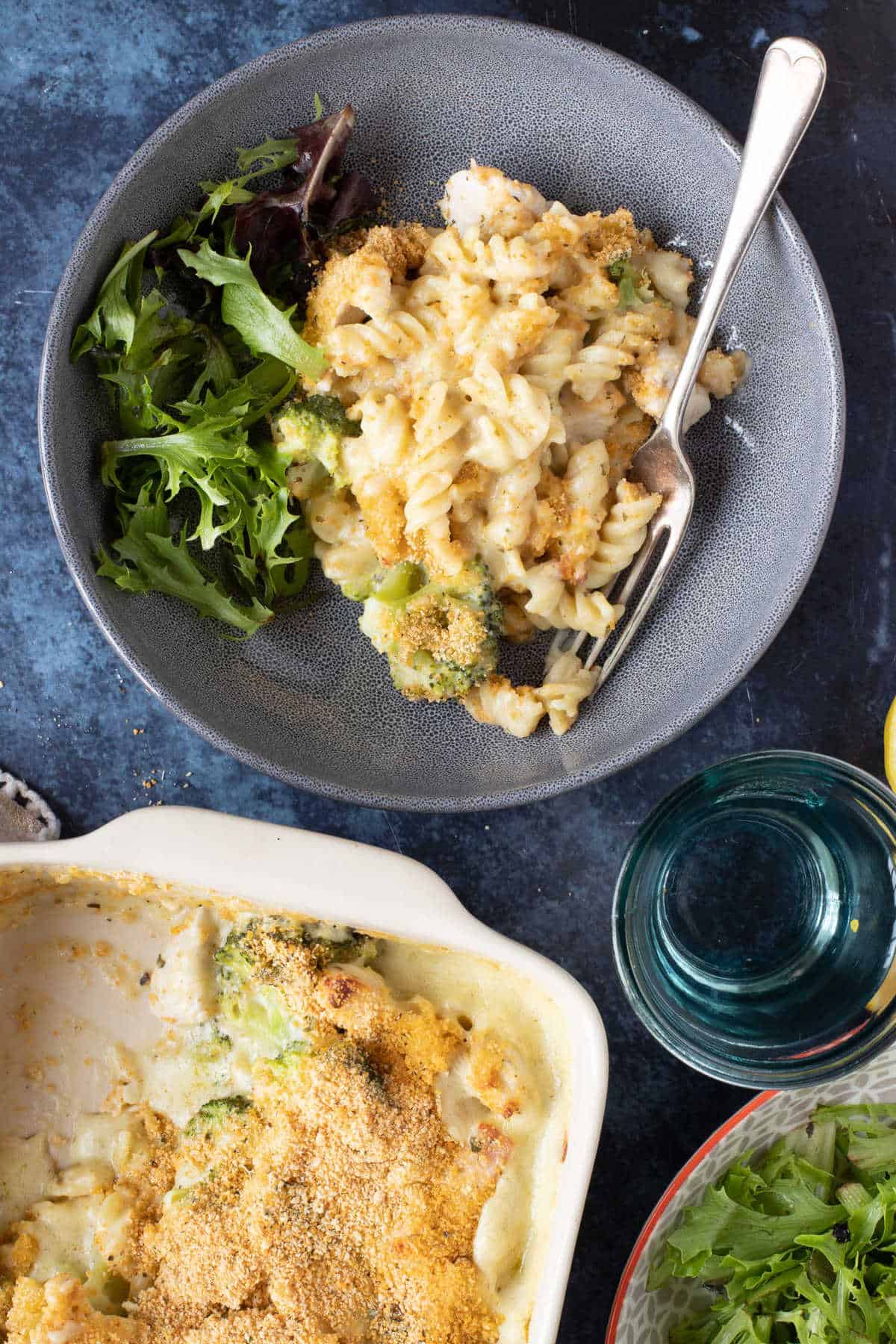 Pasta al horno en un plato gratinado con un bol de la pasta al horno y un vaso de agua.