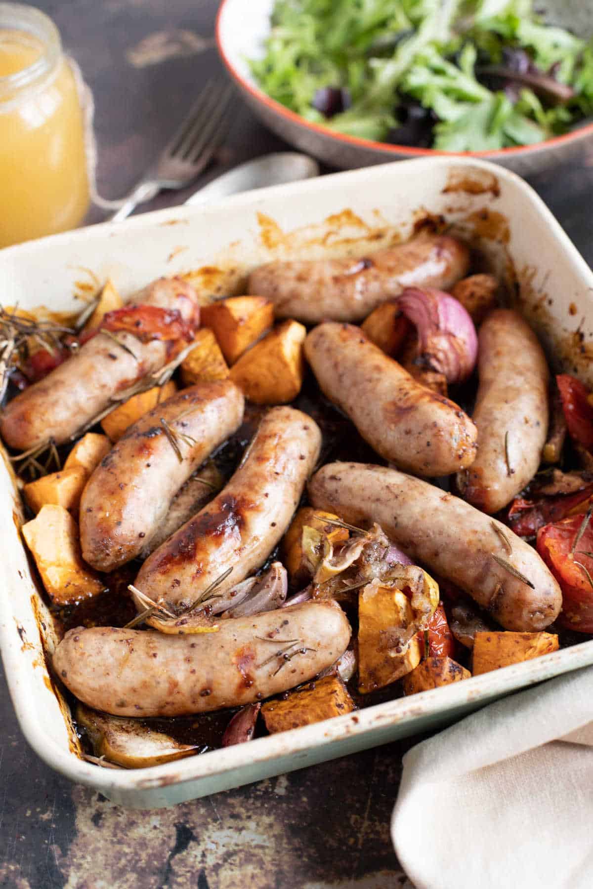 Sticky sausage traybake with seasonal vegetables in a roasting pan.