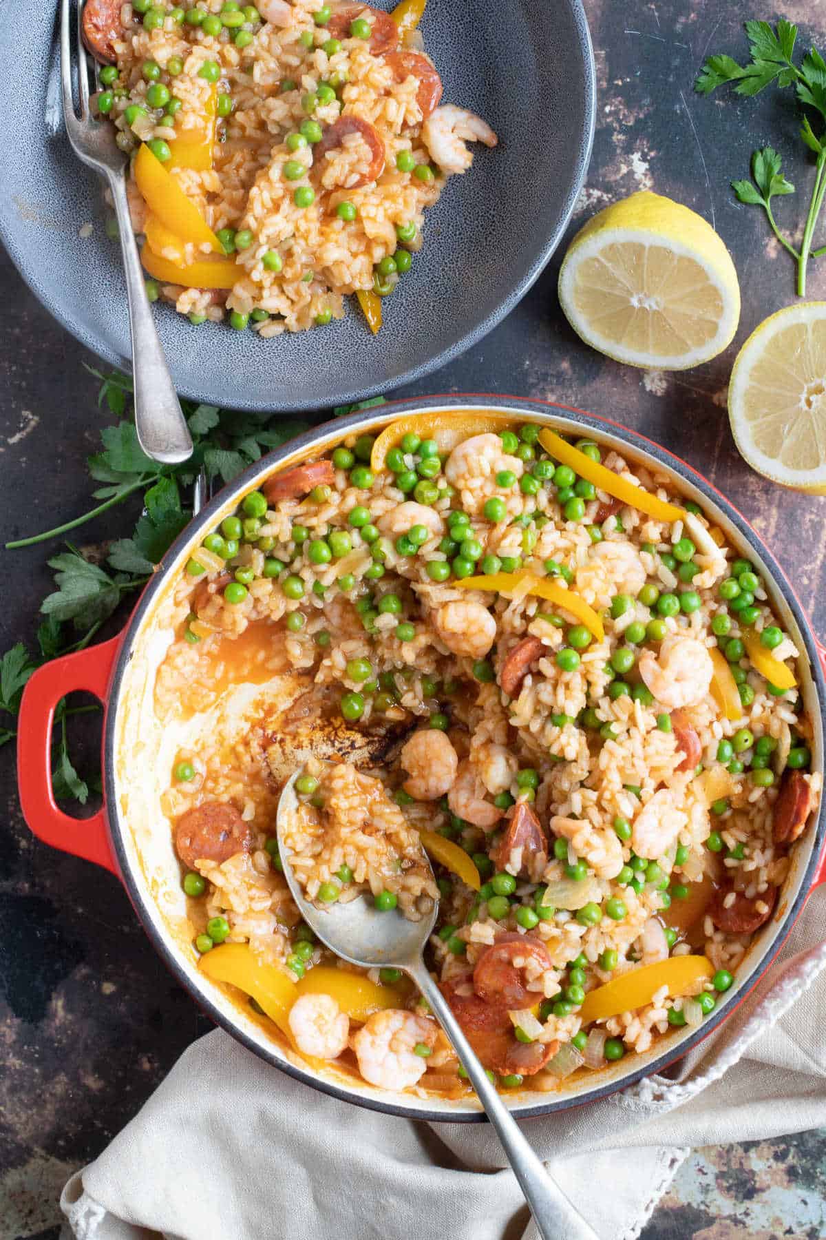 Prawn and chorizo paella in a red shallow casserole, with lemon wedges.