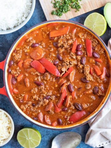 A pan of mild chilli with lime wedges, rice and tortilla chips.