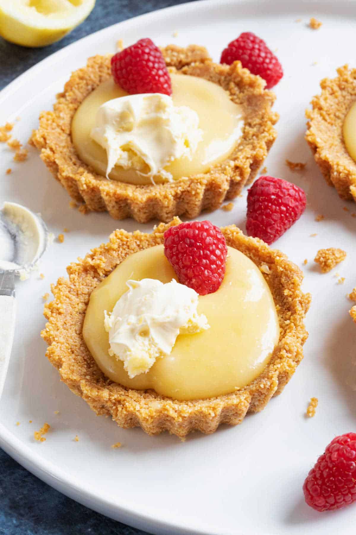 Mini lemon curd tartlets with a biscuit base, topped with raspberries and cream.