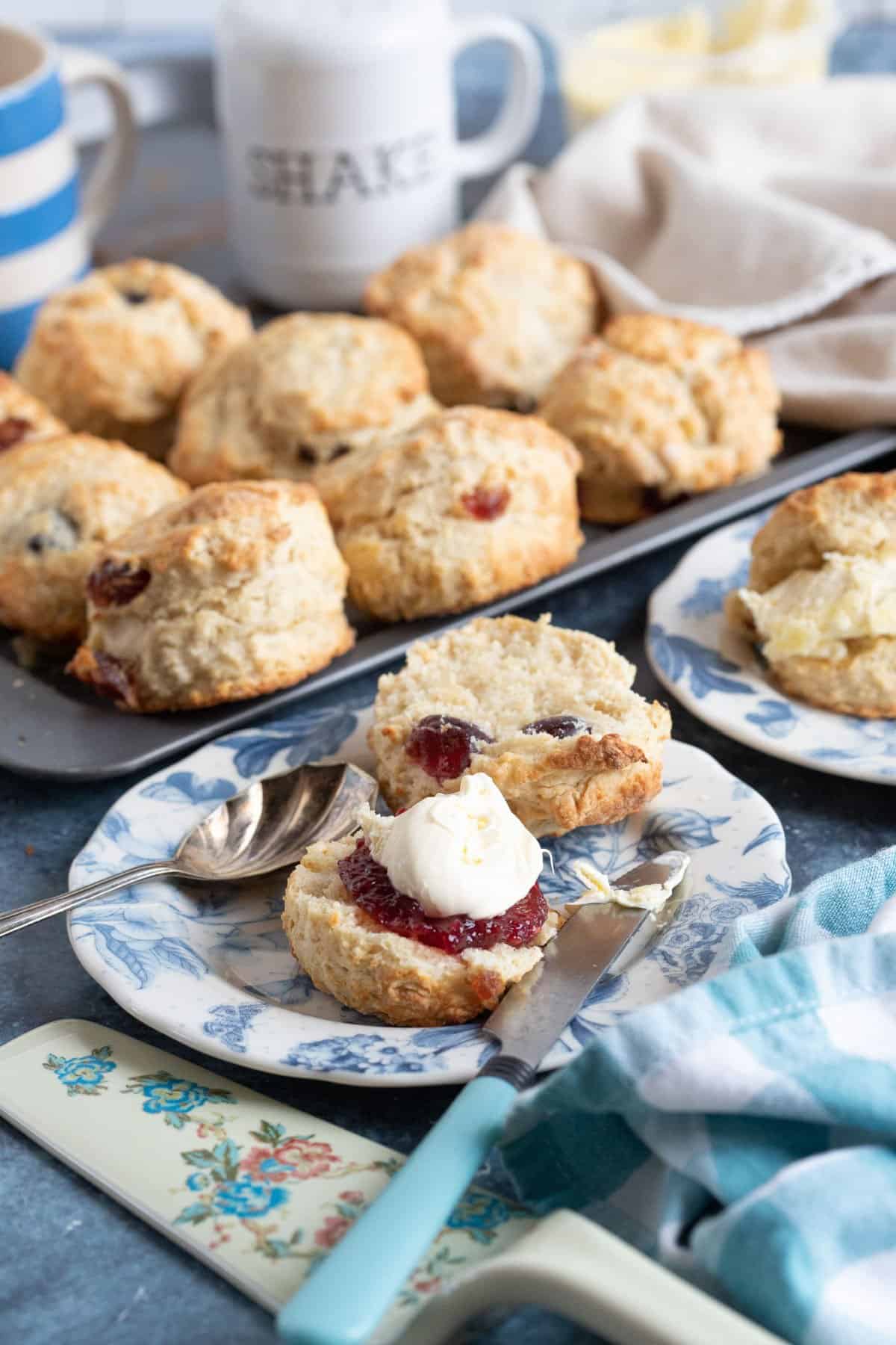 A tray of cherry and almond scones. One scone is on a plate in the foreground and split in half with jam and clotted cream.
