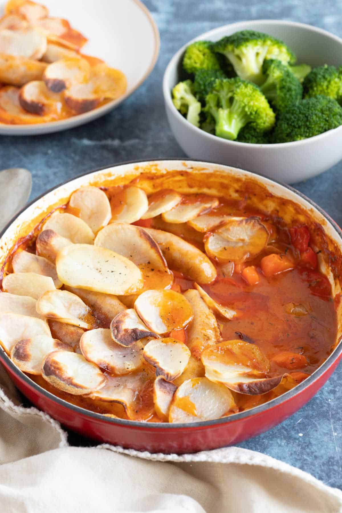Sausage hotpot in a red pan with a side of broccoli.