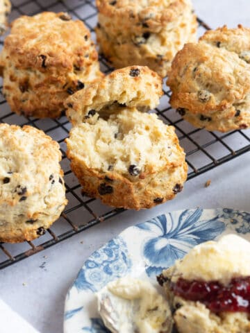 Currant scones on a cooling rack with one in the foreground split in half with jam and clotted cream.