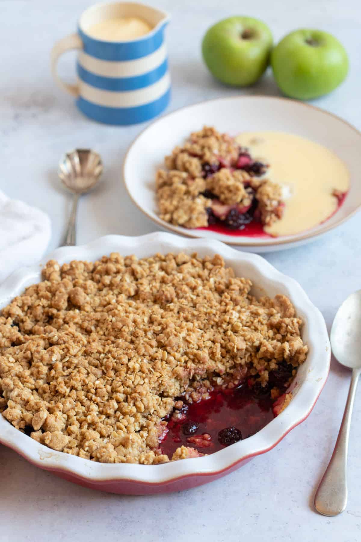 Blackberry and apple crumble in a red pie dish with a jug of custard.