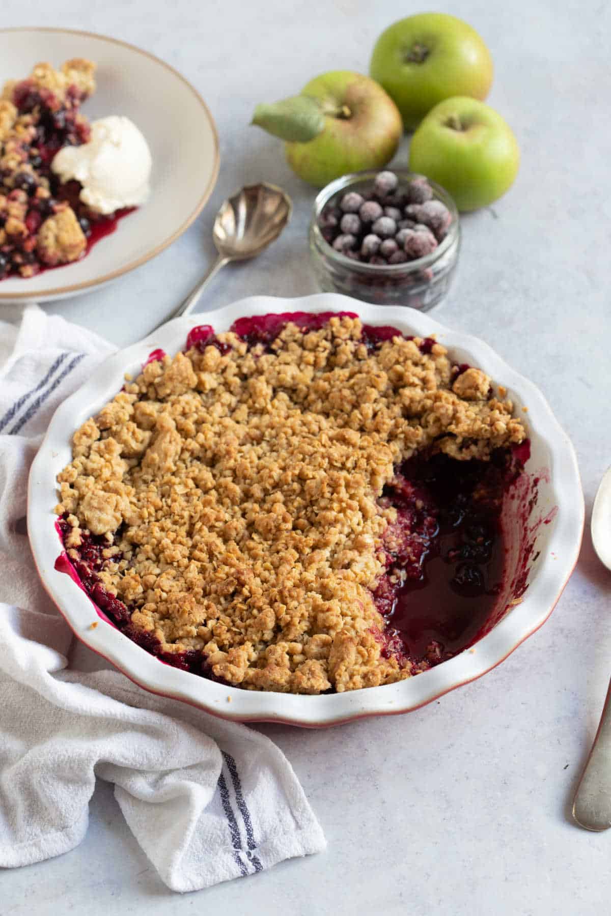 Blackcurrant crumble in a pie dish with a bowl of blackcurrants and Bramley apples behind.