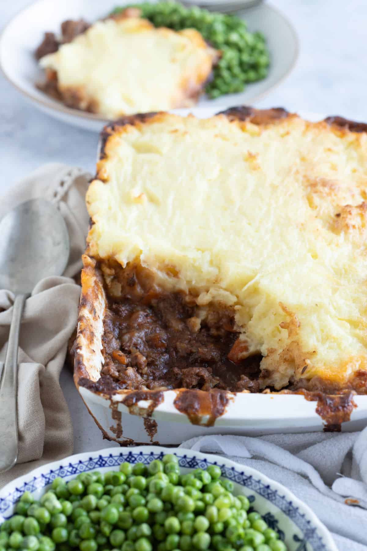 Cottage pie in a white baking dish with a bowl of peas.