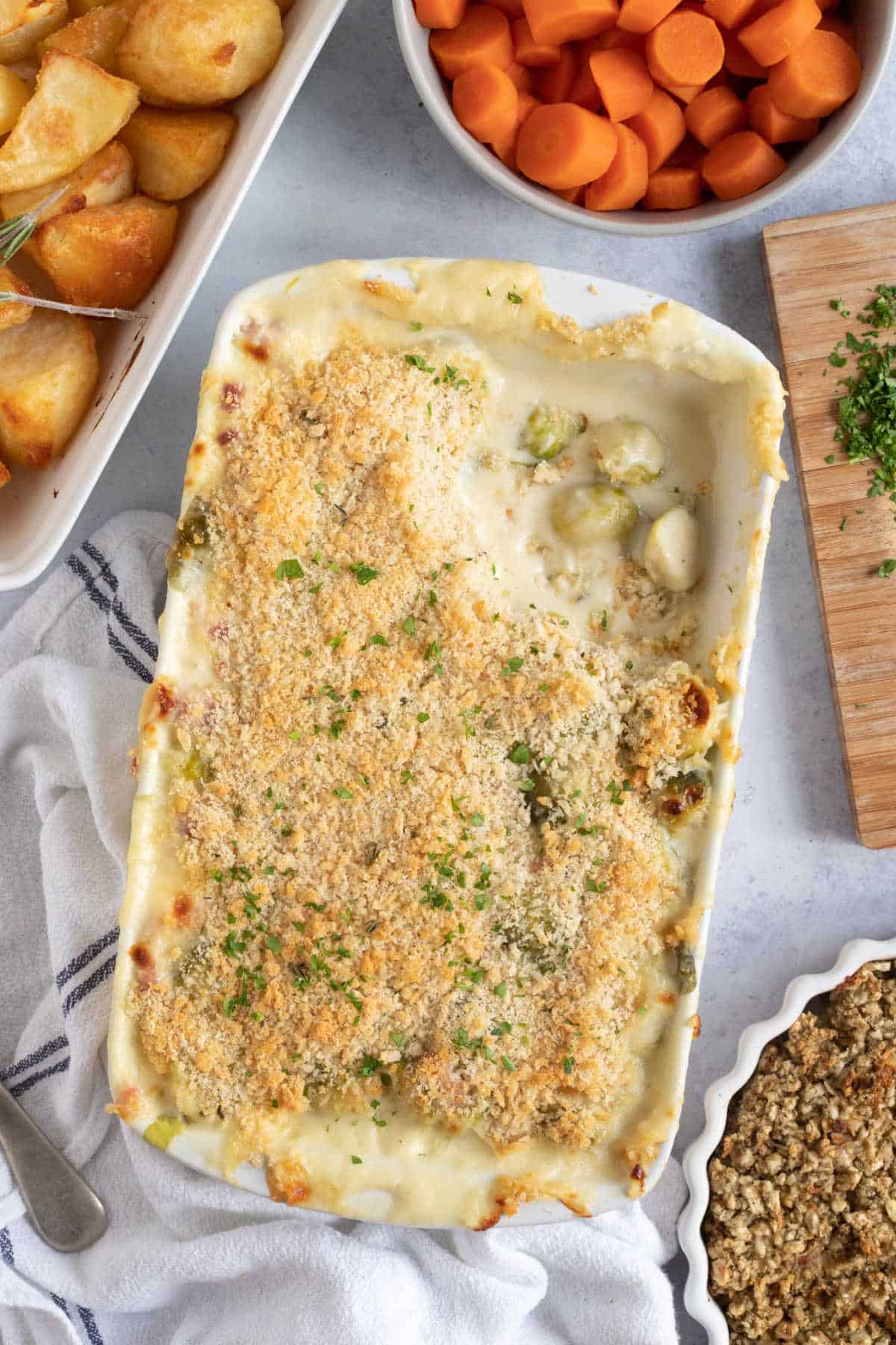 Cheesy sprout gratin in a baking dish with a bowl of carrots and roast potatoes.