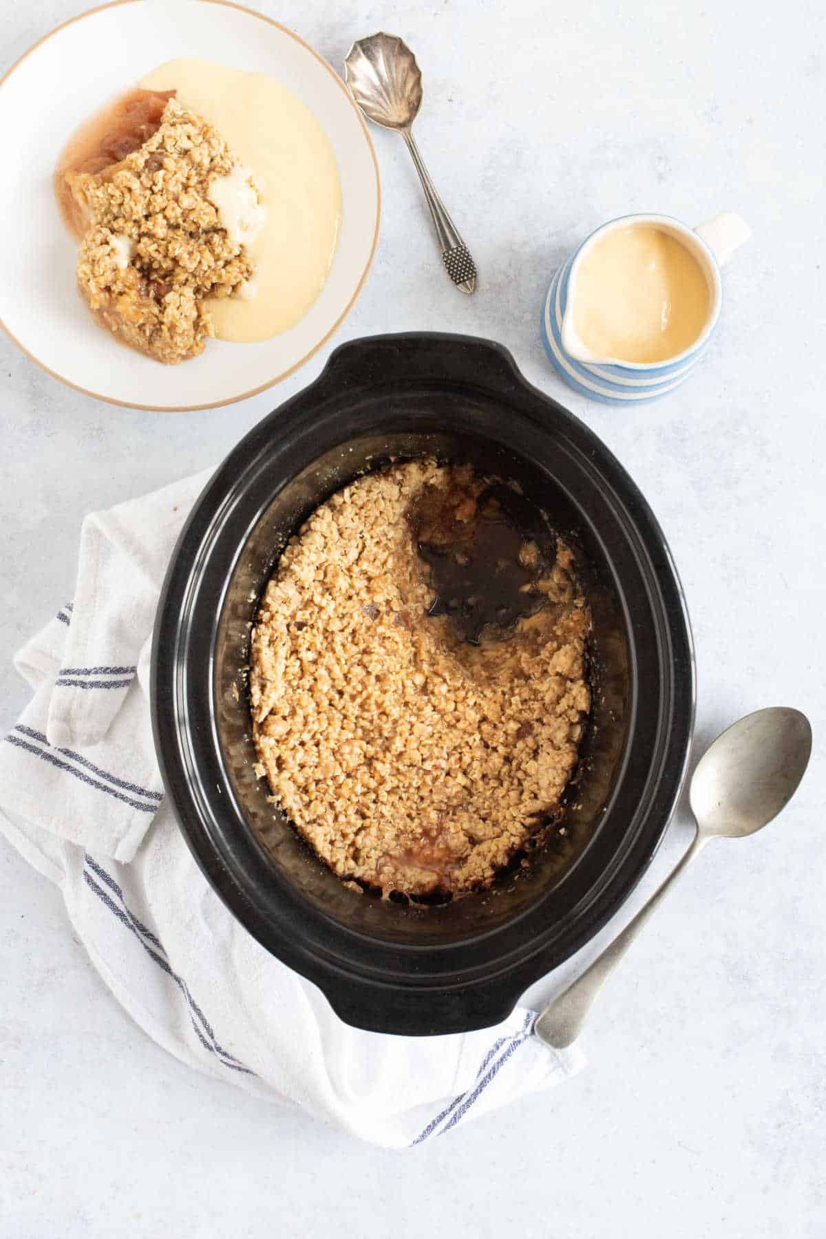 Slow cooker apple crumble in a black slow cooker basin with a bowl of apple crumble and custard on the side.