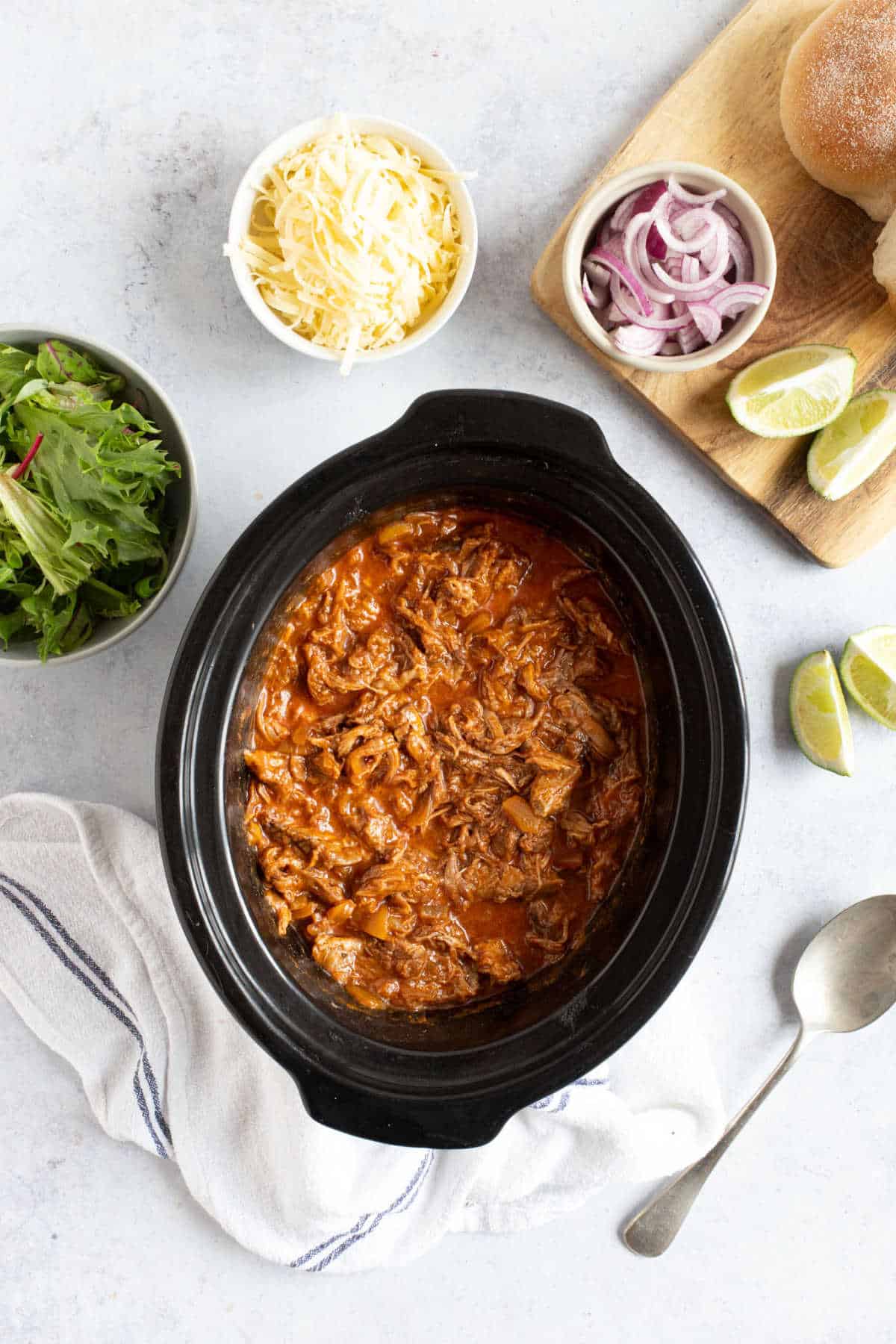 Pulled beef in a slow cooker with grated cheese, lime wedges and red onion.