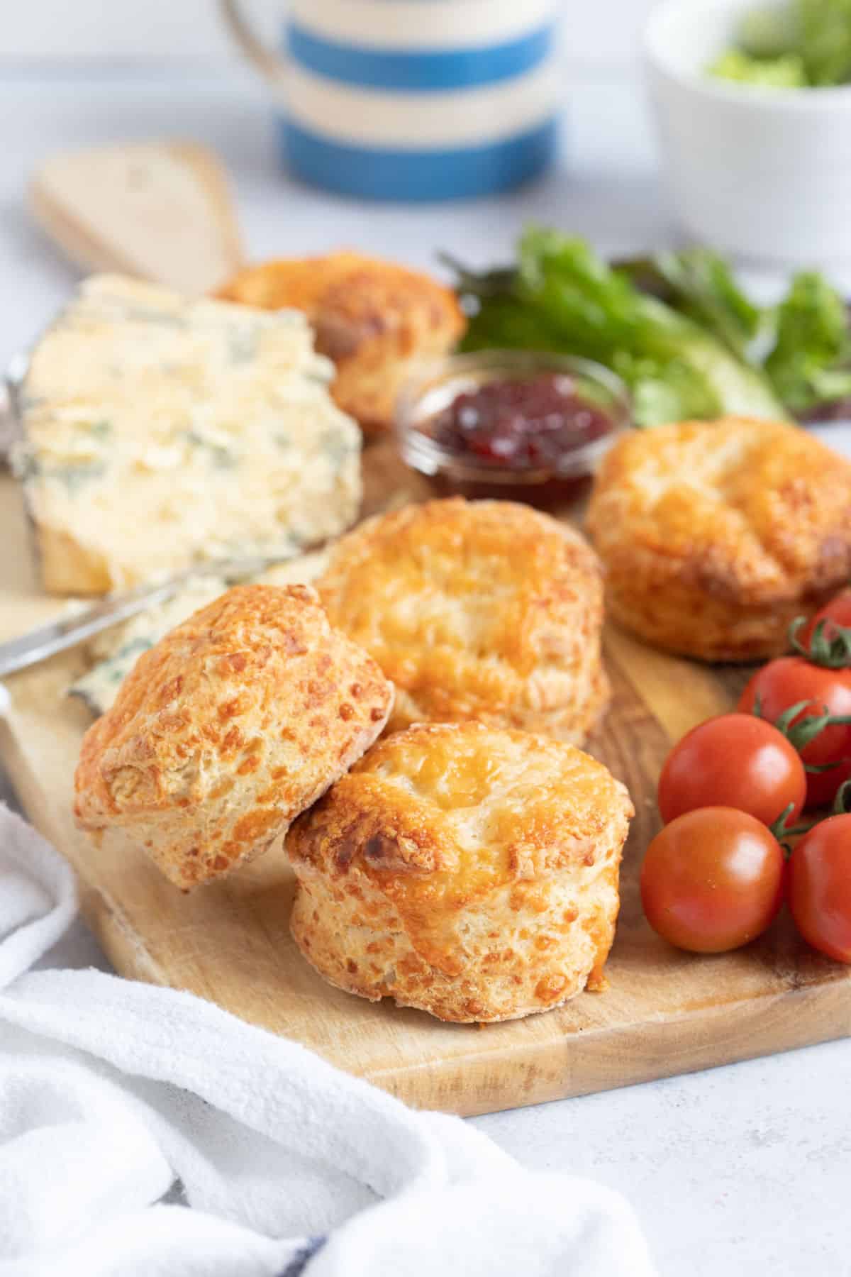 Cheese scones on a wooden serving board with cherry tomatoes, chutney, salad leaves and crumbly blue cheese.