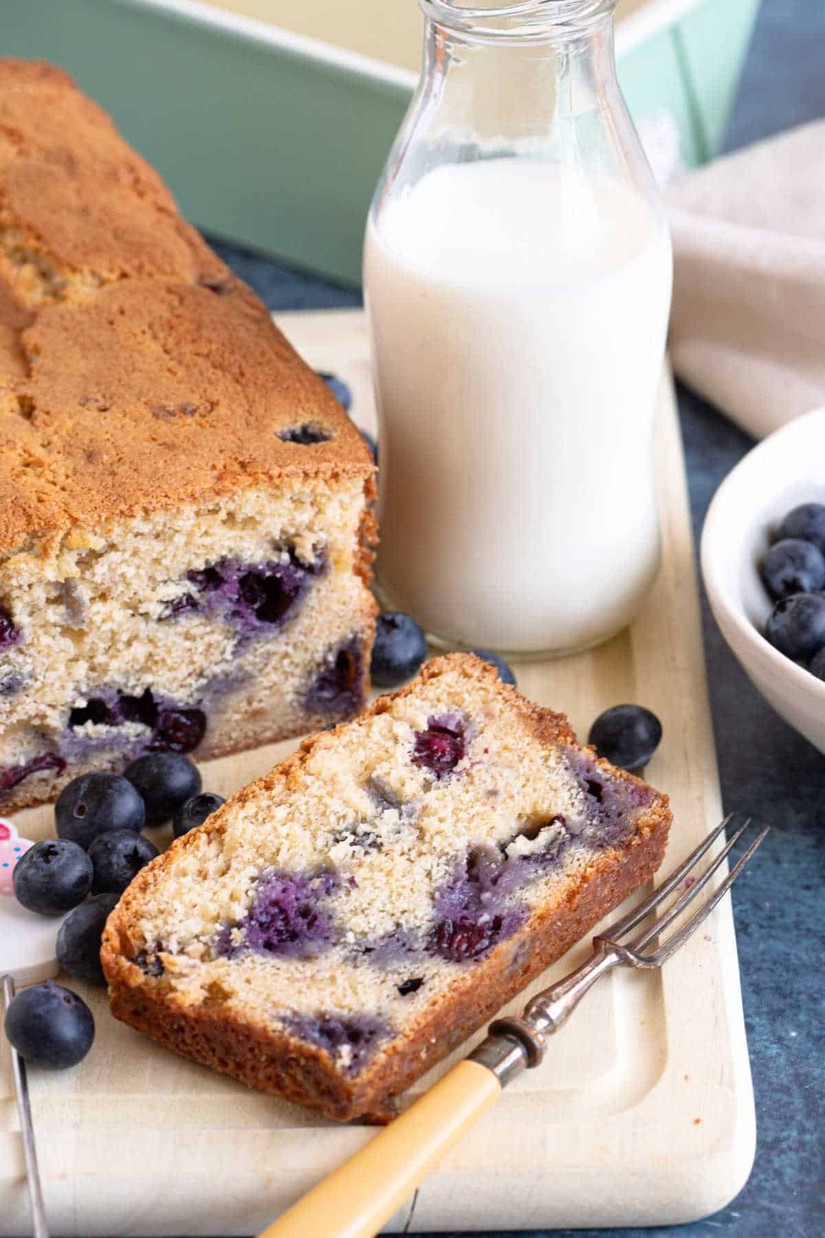 A banana and blueberry bread on a wooden board with a glass of milk behind.