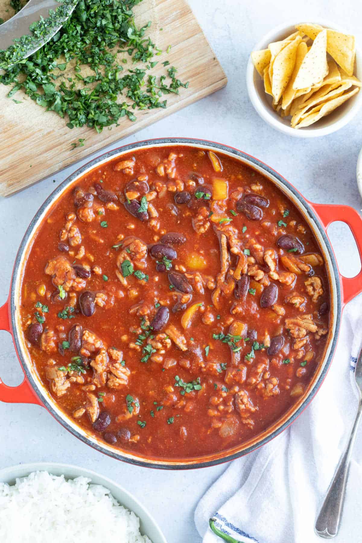 Turkey mince chilli in a red pan with chopped coriander and tortillas on the side.