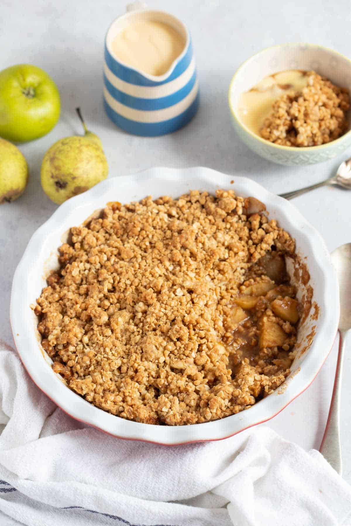 Apple and pear crumble in a red pie dish wit ha jug of custard next to it.