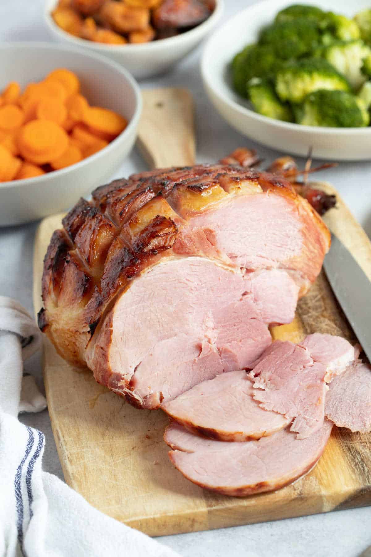 Slow cooked gammon joint with a honey mustard glaze on a wooden board with carrots, broccoli, and roasted sweet potatoes in the background.