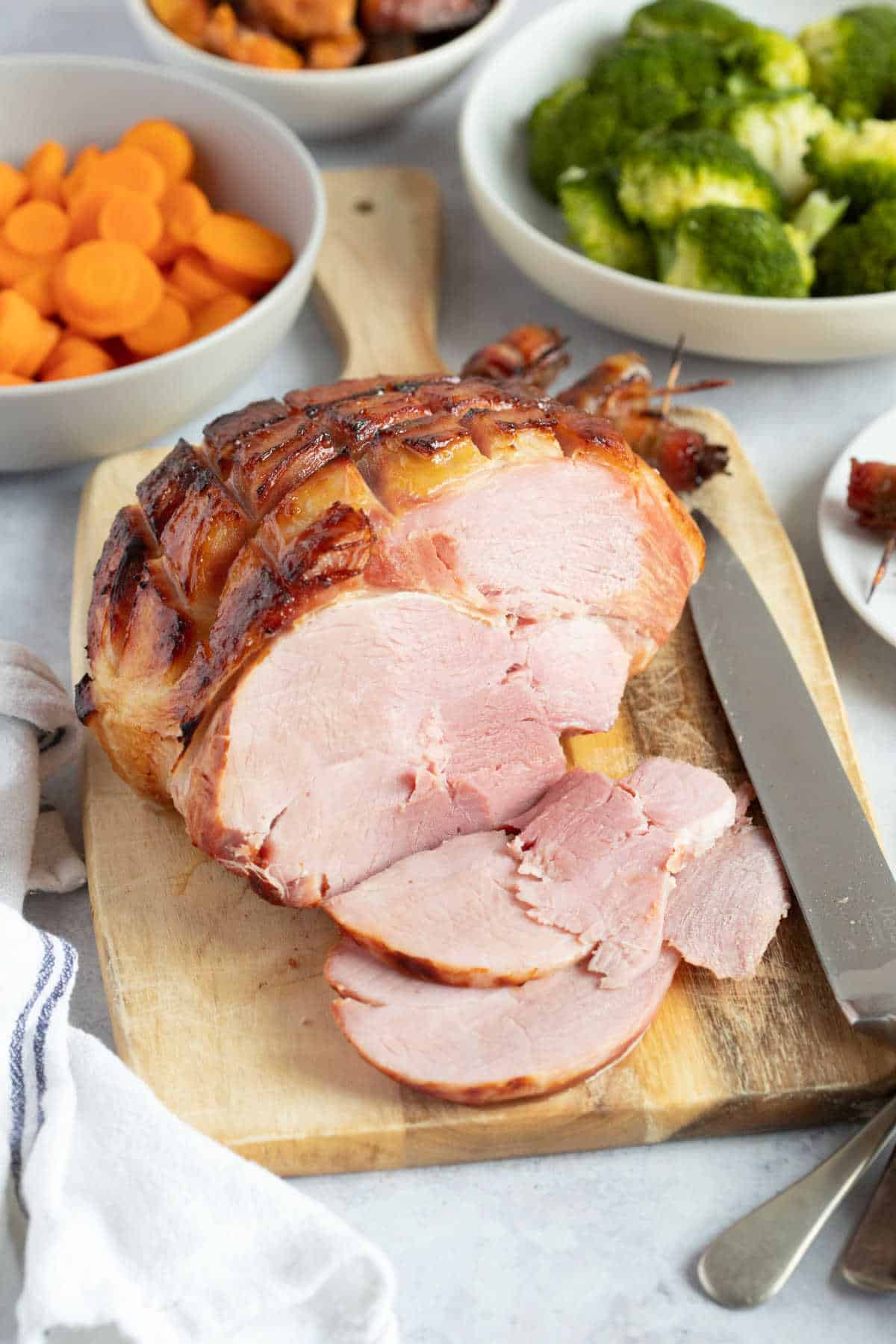 Cooked gammon joint on a wooden board with bowls of vegetables in the background.