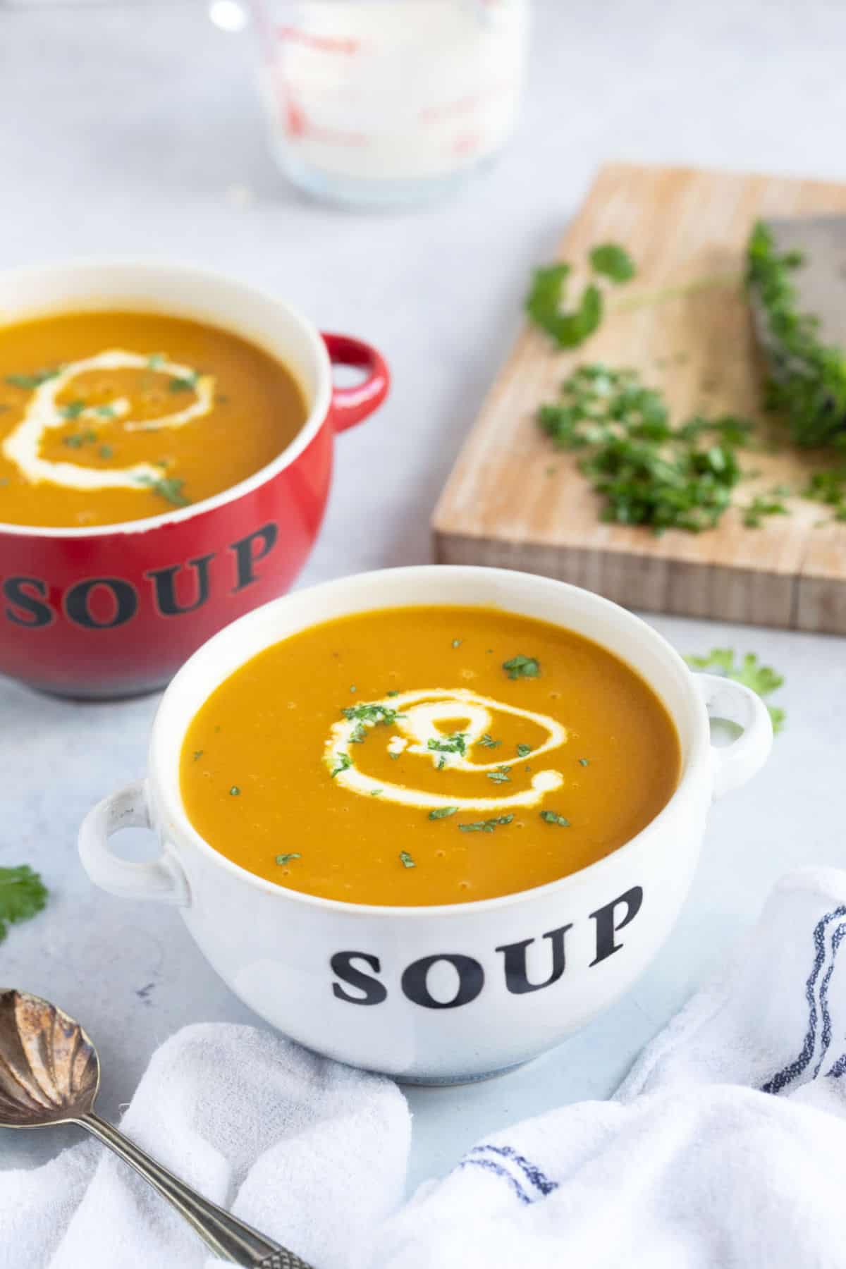Two bowls of spiced carrot and parsnip soup with a jug of cream in the background.